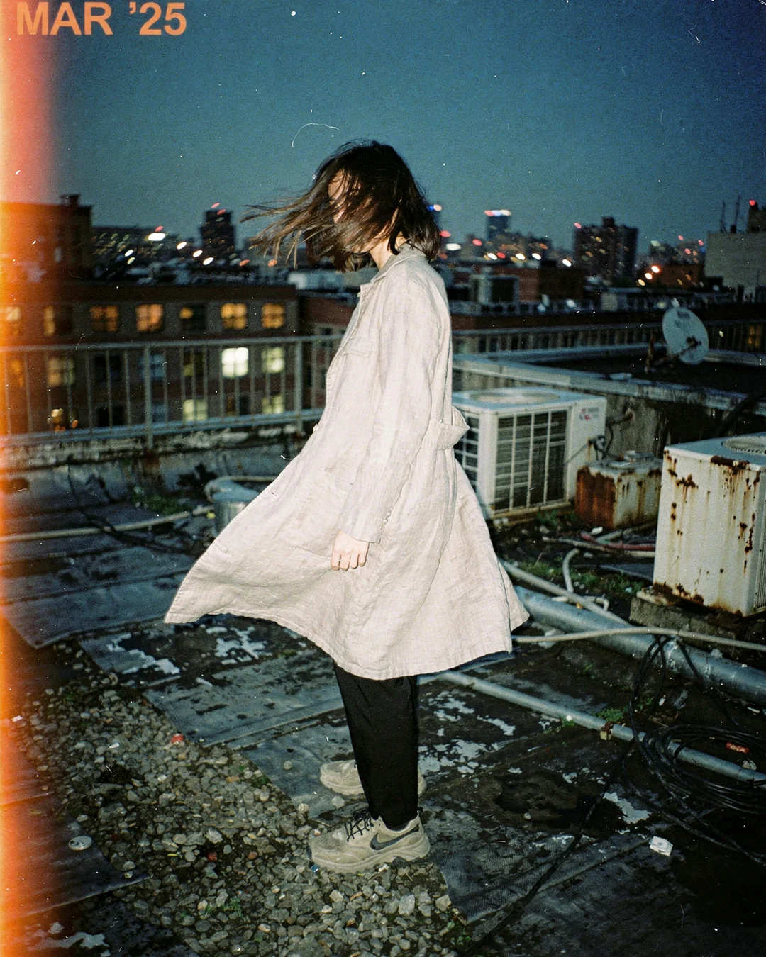 Rooftop fashion editorial at blue hour with wind-blown coat and city backdrop, comparing product photography and AI virtual try-on