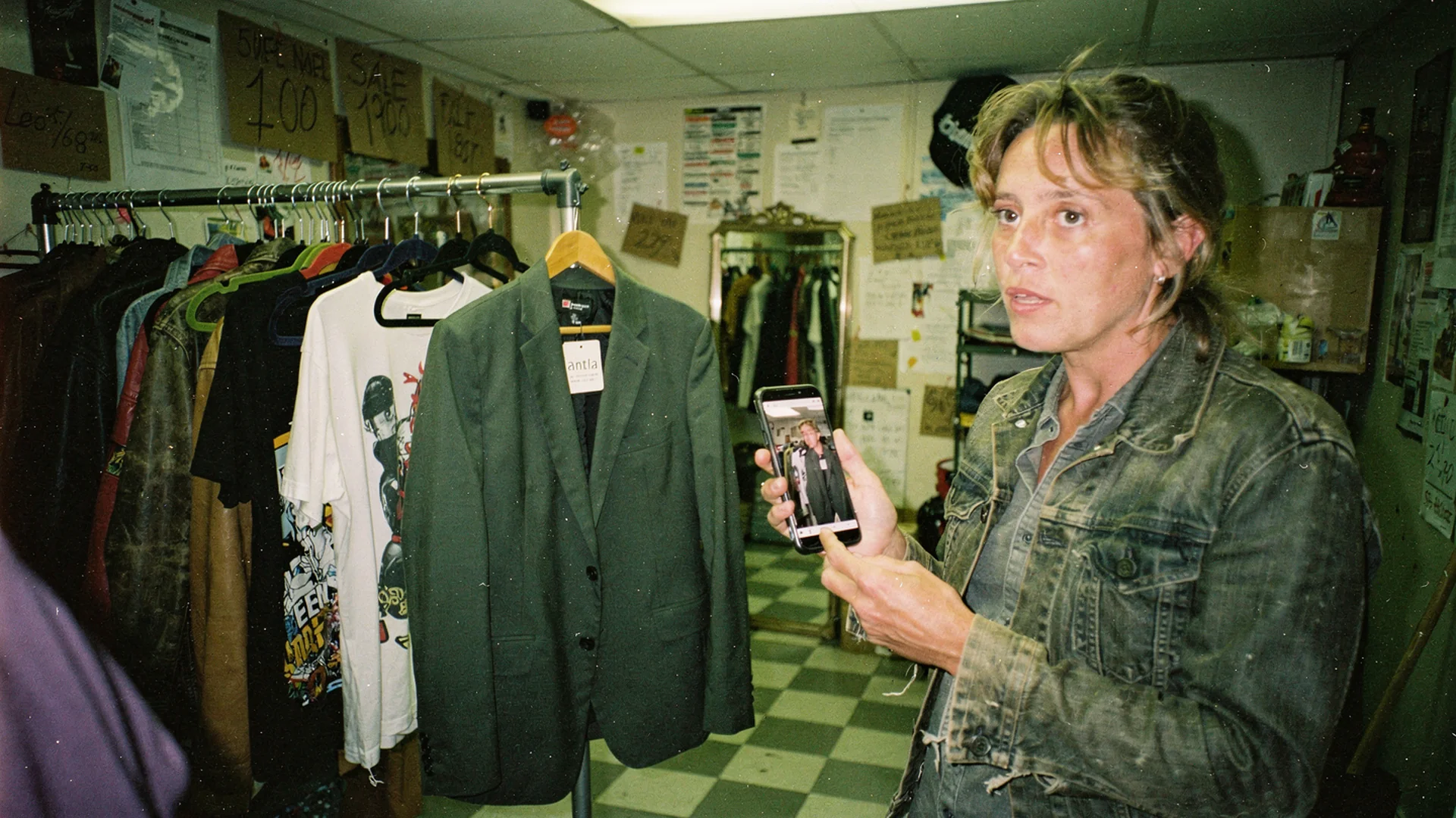 Independent clothing store back room with rolling rack and owner previewing blazer try-on on phone