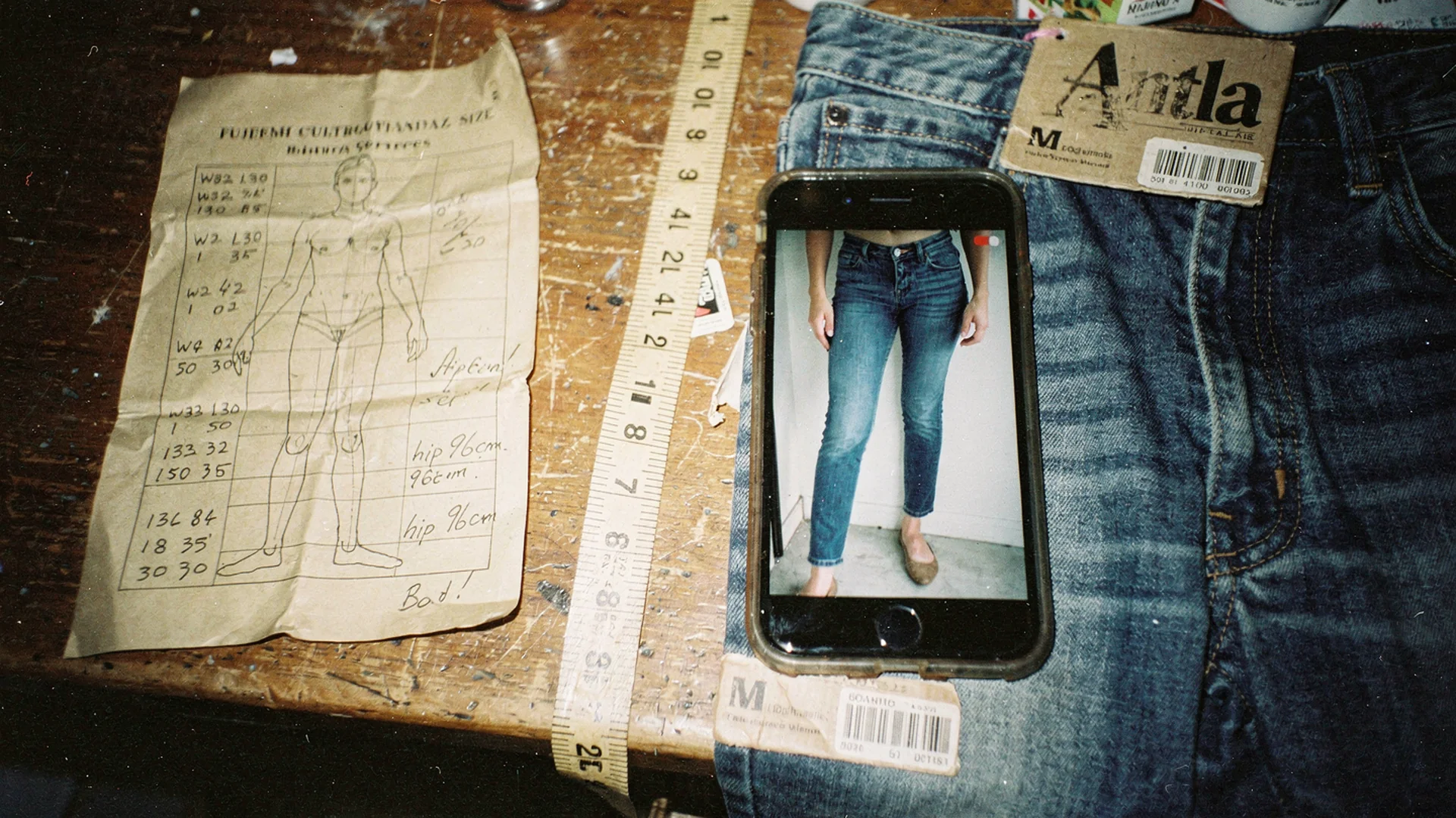 Crumpled size chart next to phone showing mirror try-on preview of jeans on a worn table