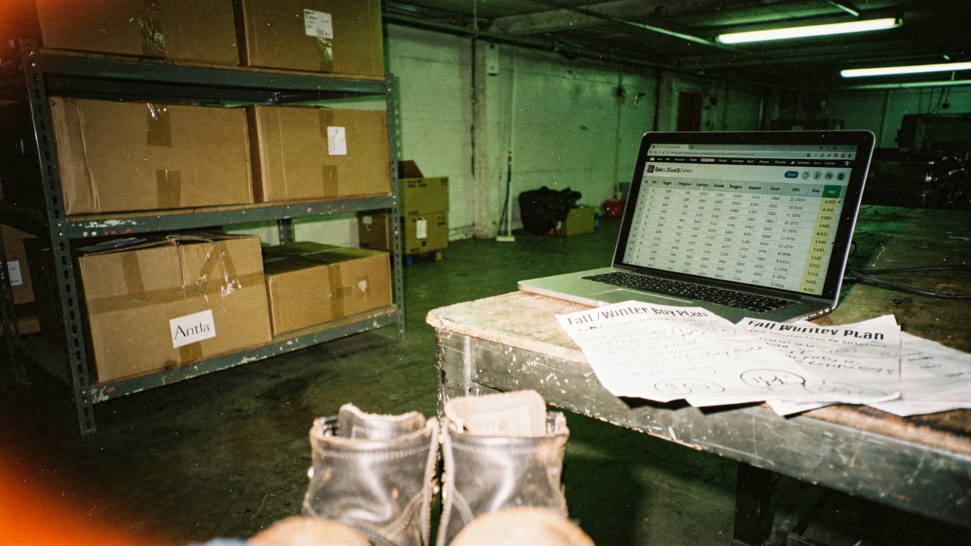Warehouse shelf of fashion cartons beside a demand forecast spreadsheet and seasonal buy plan on a tablet