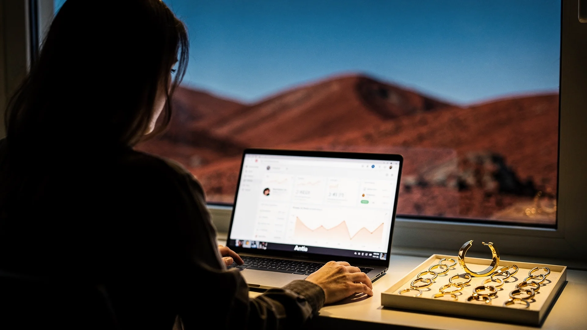 Jewelry merchandiser reviewing Try-on feed analytics on laptop with earring and necklace samples on desk in natural light