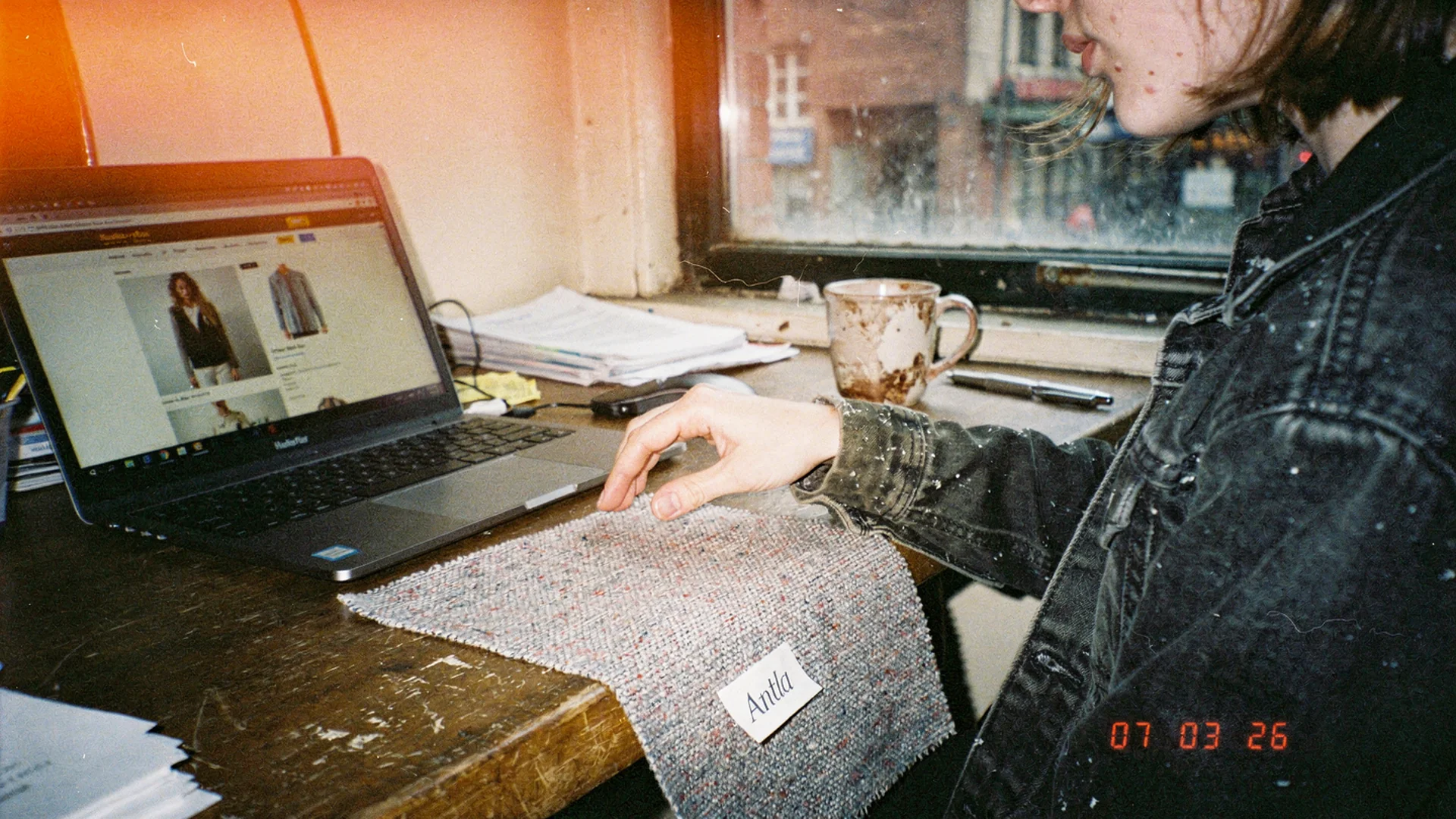 Close editorial of hands near fabric swatches beside a laptop showing an online dress product page