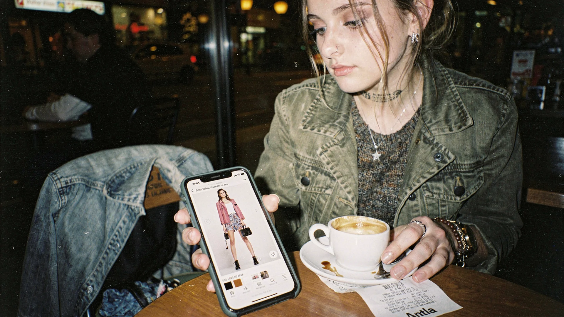 Fashion shopper absorbed in a mobile product page virtual try-on at a cafe table late in the evening