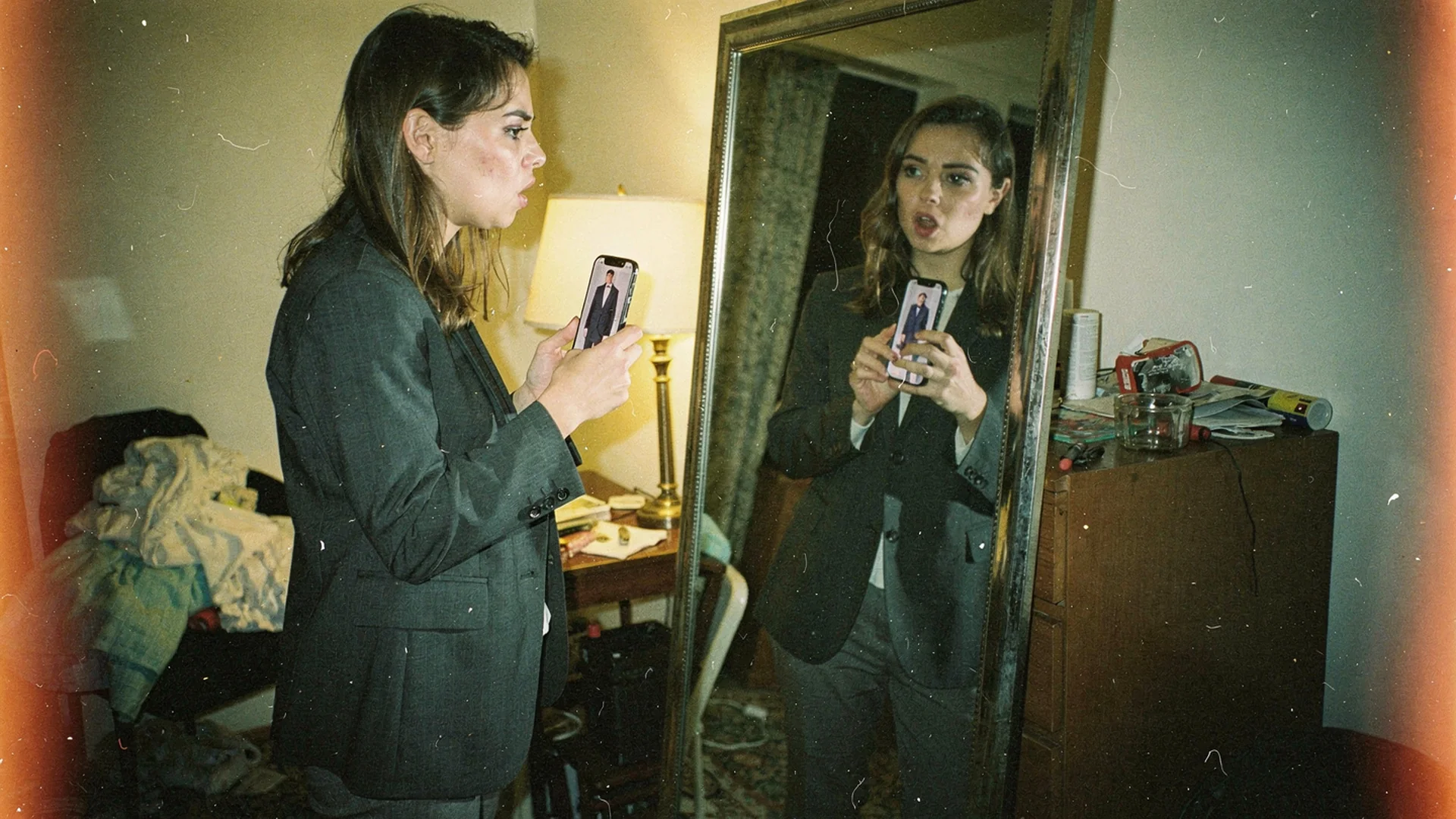 Shopper comparing a phone virtual try-on preview of a blazer against her own reflection in an apartment mirror