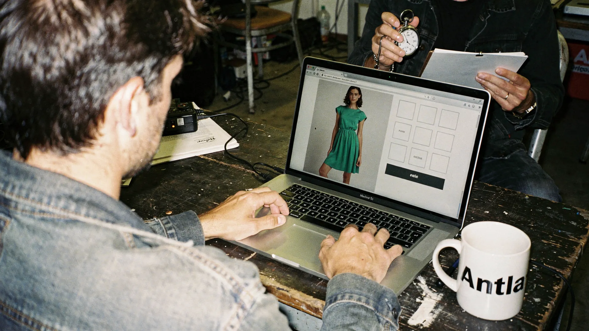 Usability session with a shopper at a laptop showing a green dress and size grid, a researcher with a stopwatch, an Antla mug