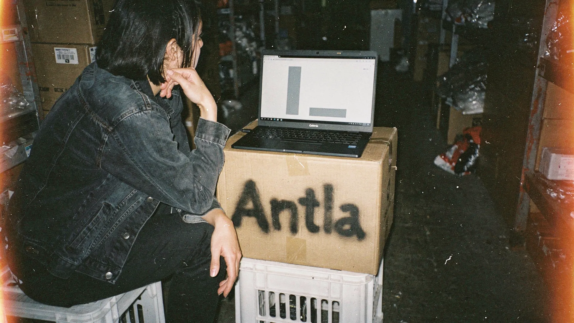 Fashion merchandiser in a stockroom studying a laptop chart with one tall bar and one short bar, set on a box stenciled Antla