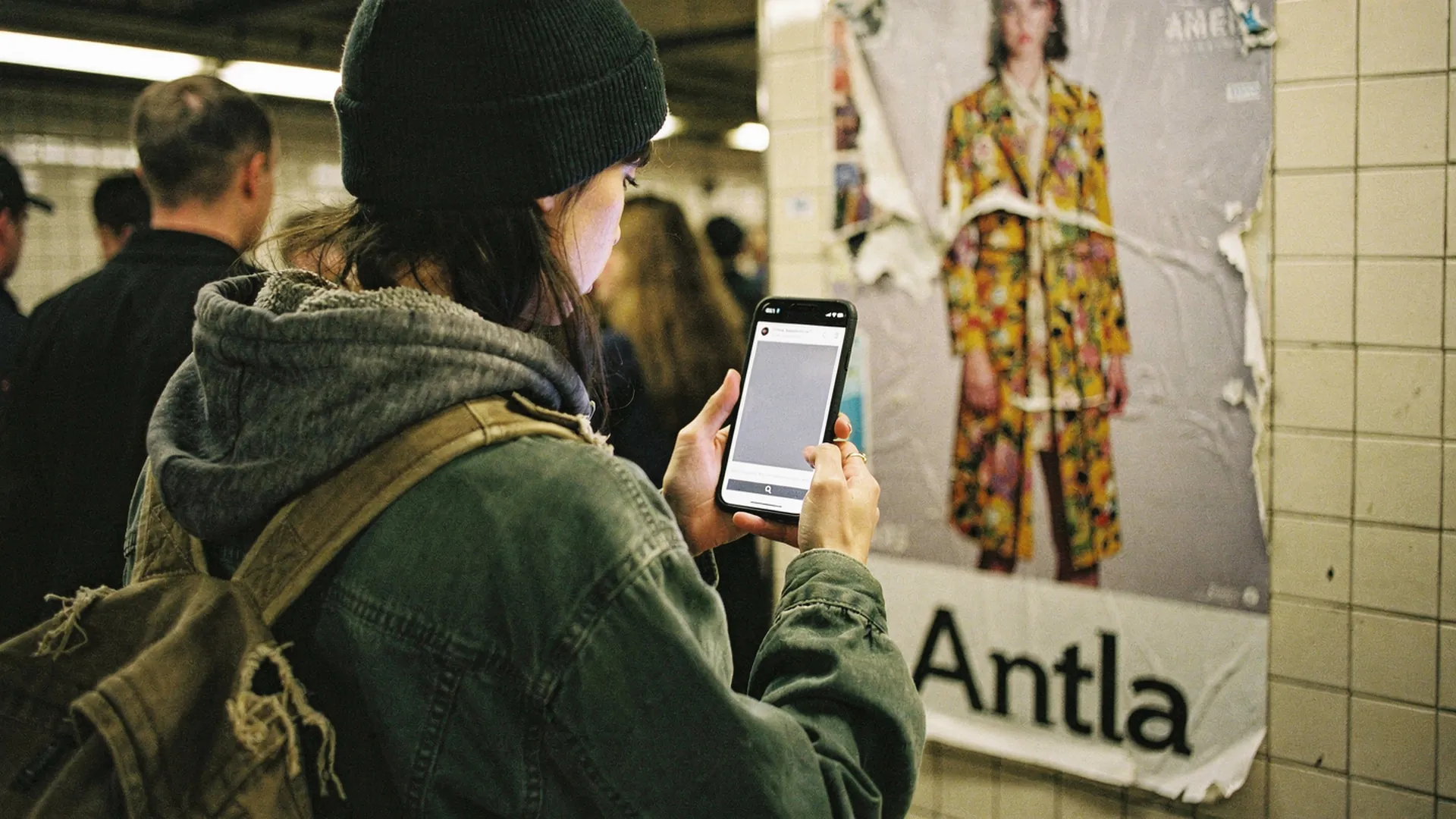Commuter on a subway platform staring at a phone stuck on a grey loading screen beside a torn Antla fashion poster