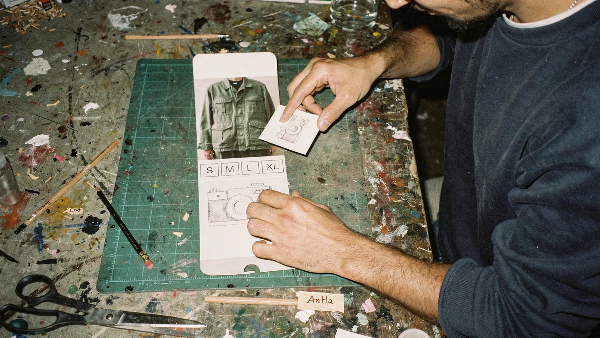Paper mockups of a phone product page on a cutting mat, with a small camera-icon card moved in the column