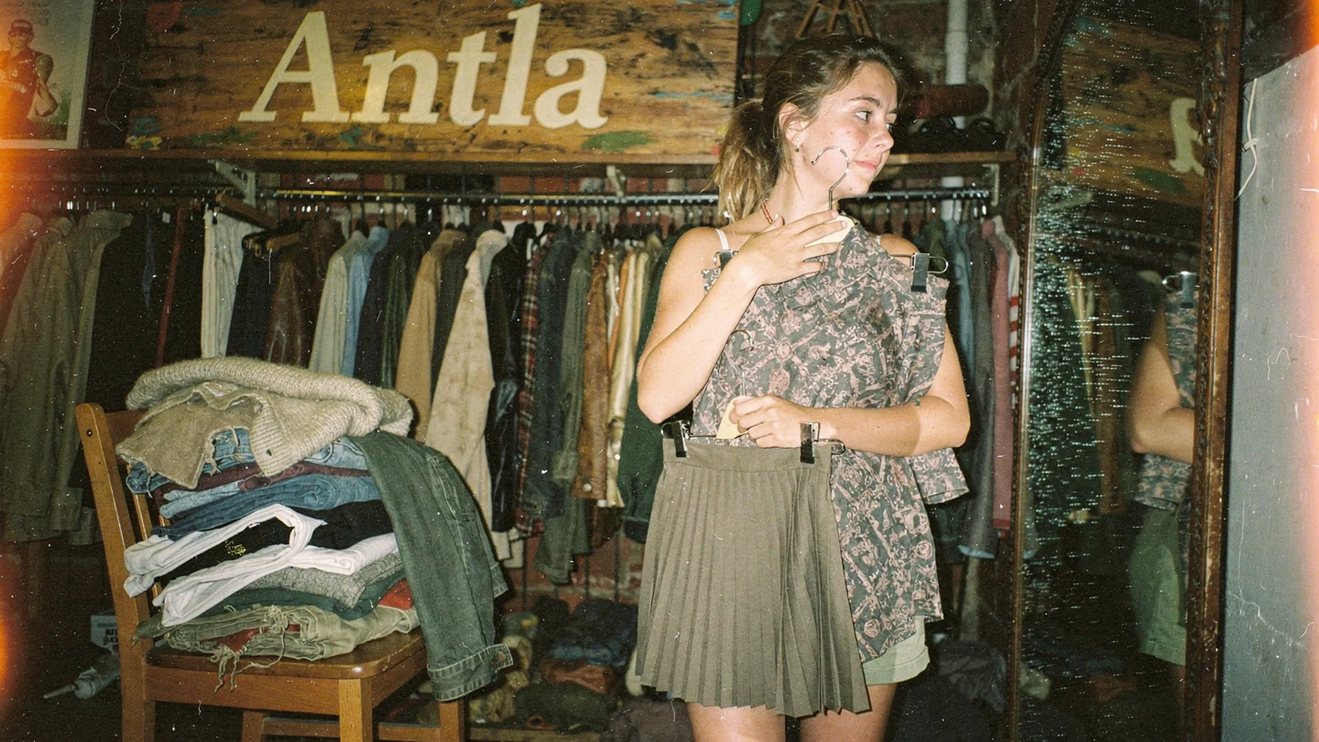 Shopper in a vintage store holding a patterned top against a pleated skirt, a chair piled with clothes, Antla sign above